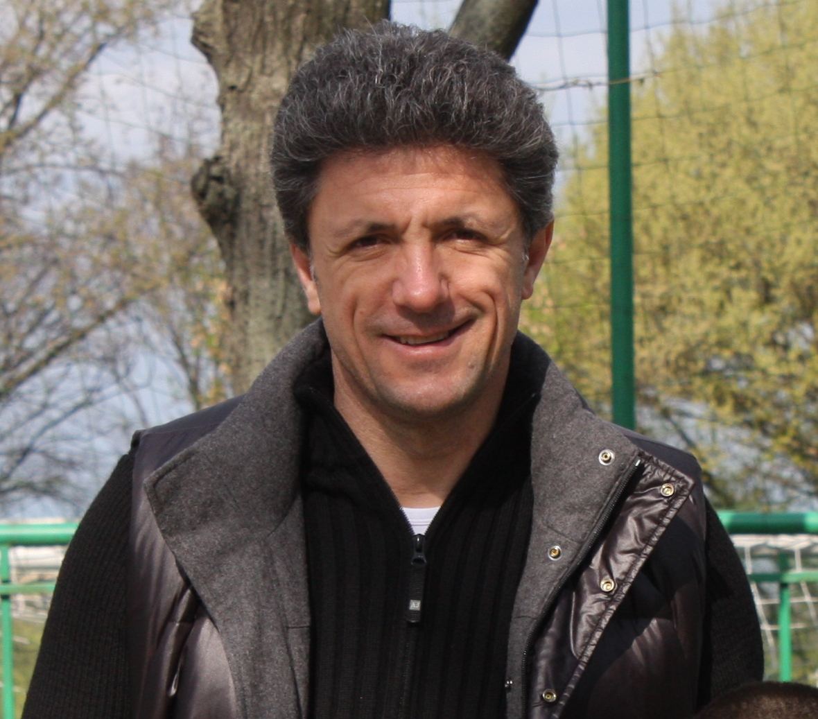 Portretul lui Gică Popescu, zâmbind, într-o jachetă întunecată, cu un gard verde și copaci în fundal.
