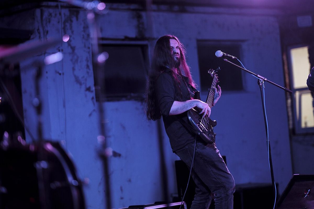 Un muzician cântă la chitară bas pe scenă, concentrat, sub lumini violet, într-o atmosferă energică de concert în seară.