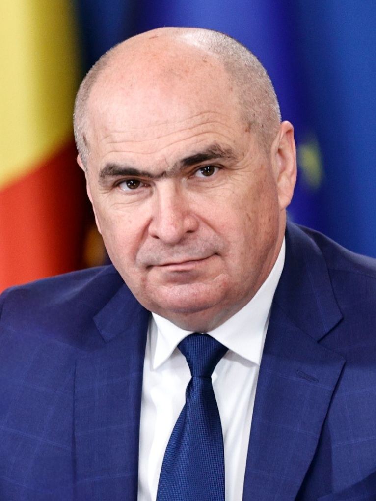 Ilie Bolojan, premierul României, în fața steagurilor României și Uniunii Europene, într-un costum albastru și cravată albastră.