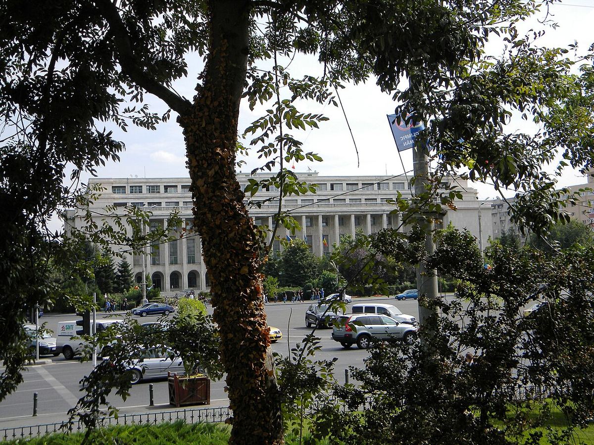 Palatul Victoria din București, sediul Guvernului României, văzut printre copaci din Piața Victoriei, pe cer cu nori.
