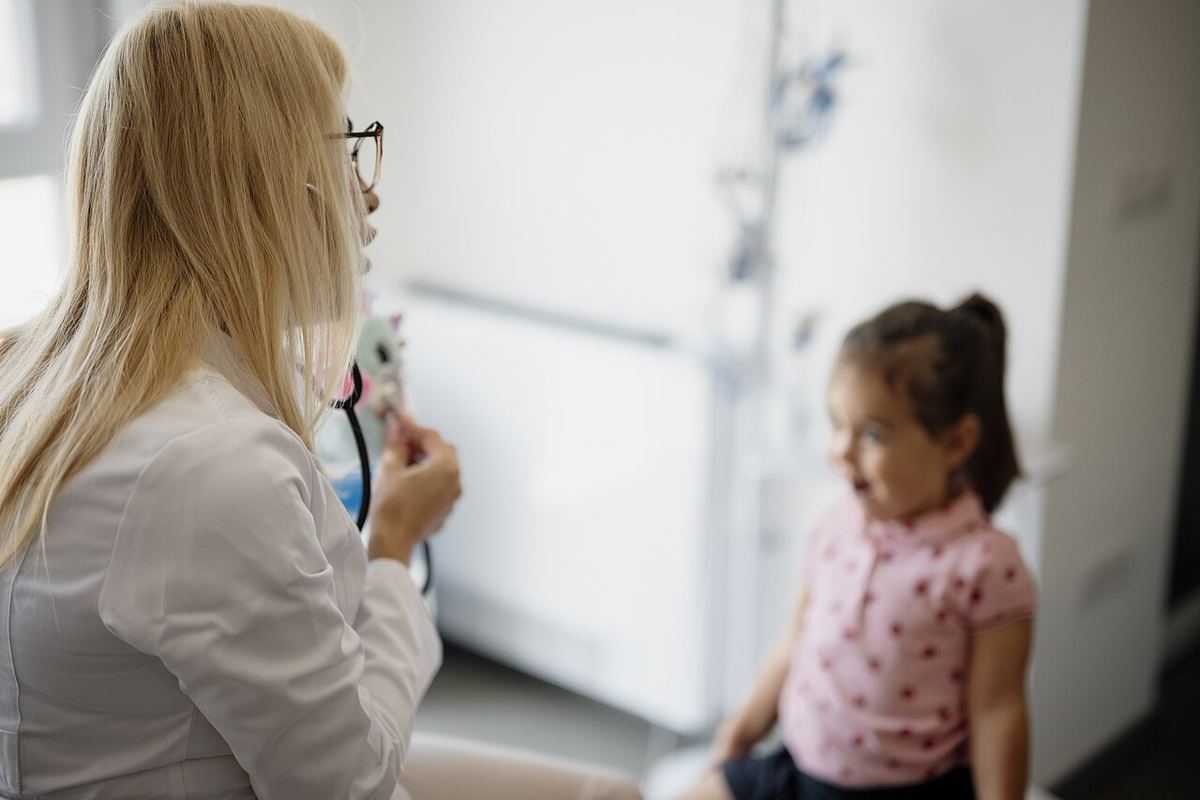Un medic în halat alb, cu ochelari, discută cu o fetiță într-un cabinet medical luminos.