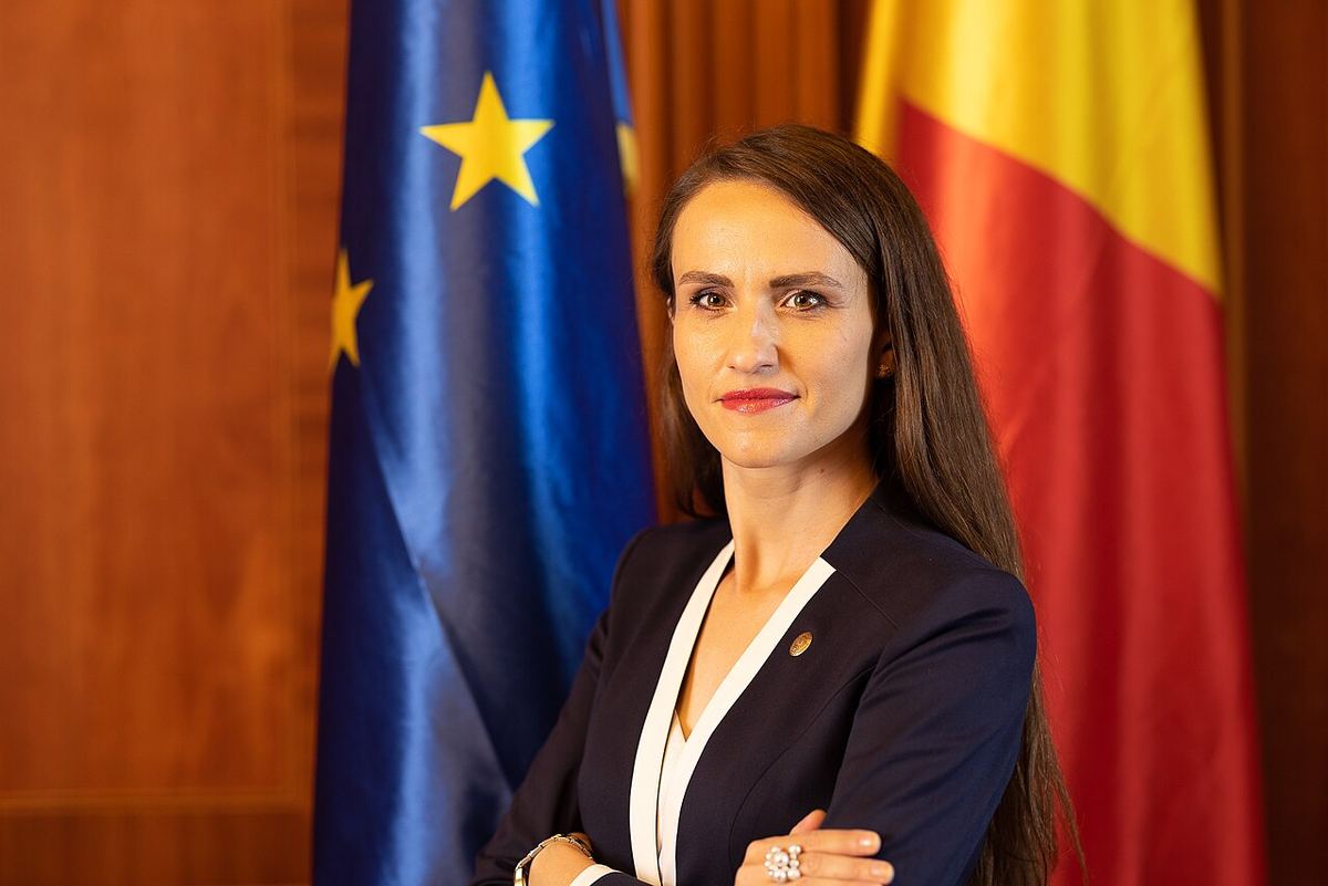 Oana Țoiu, ministra de Externe, în fața drapelelor României și Uniunii Europene, cu brațele încrucișate.