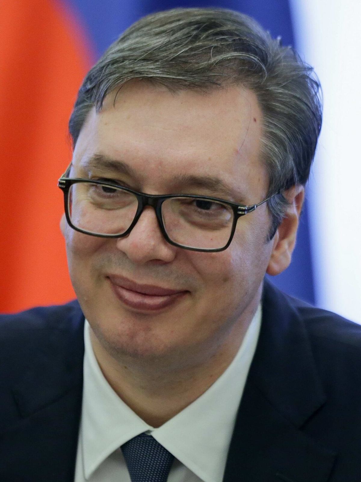 Portret de arhivă al președintelui Serbiei, Aleksandar Vučić, purtând ochelari și costum închis la culoare, în fața unui steag.