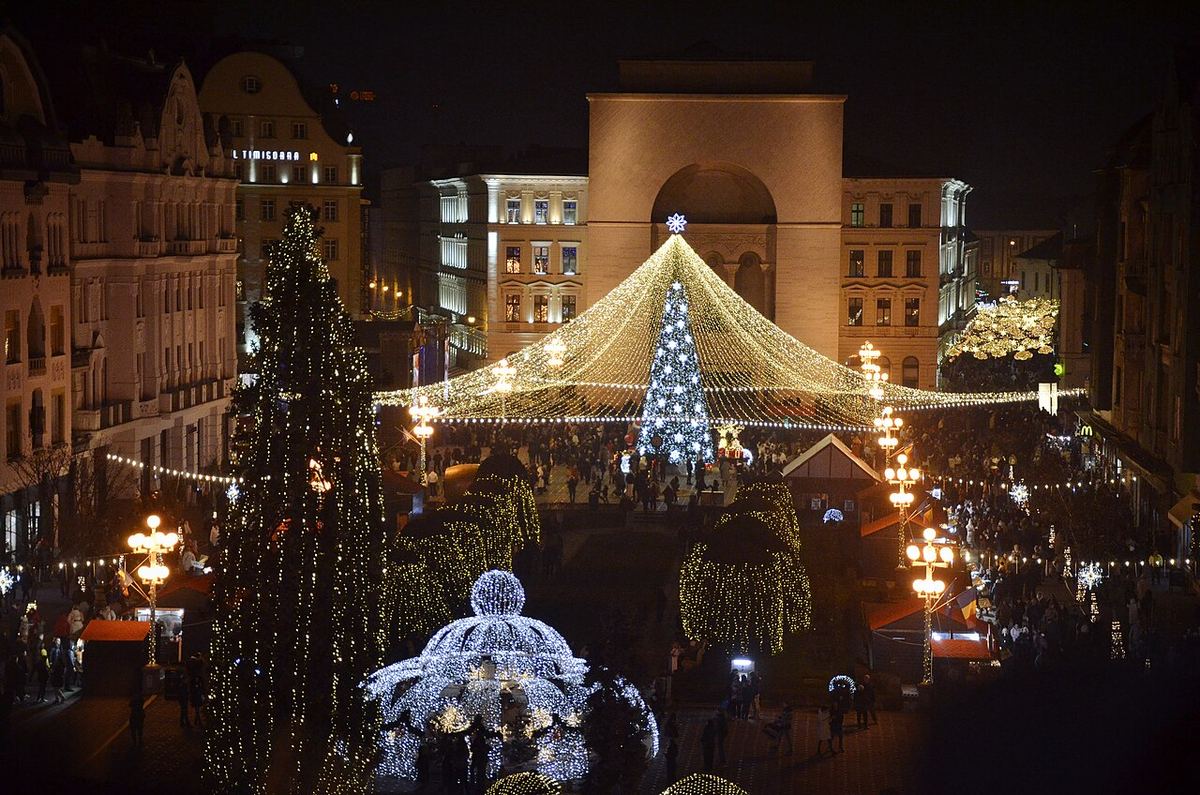 Vedere nocturnă de sus a târgului de Crăciun din Piața Victoriei, Timișoara, cu iluminat festiv strălucitor și clădirea Operei Naționale în fundal.