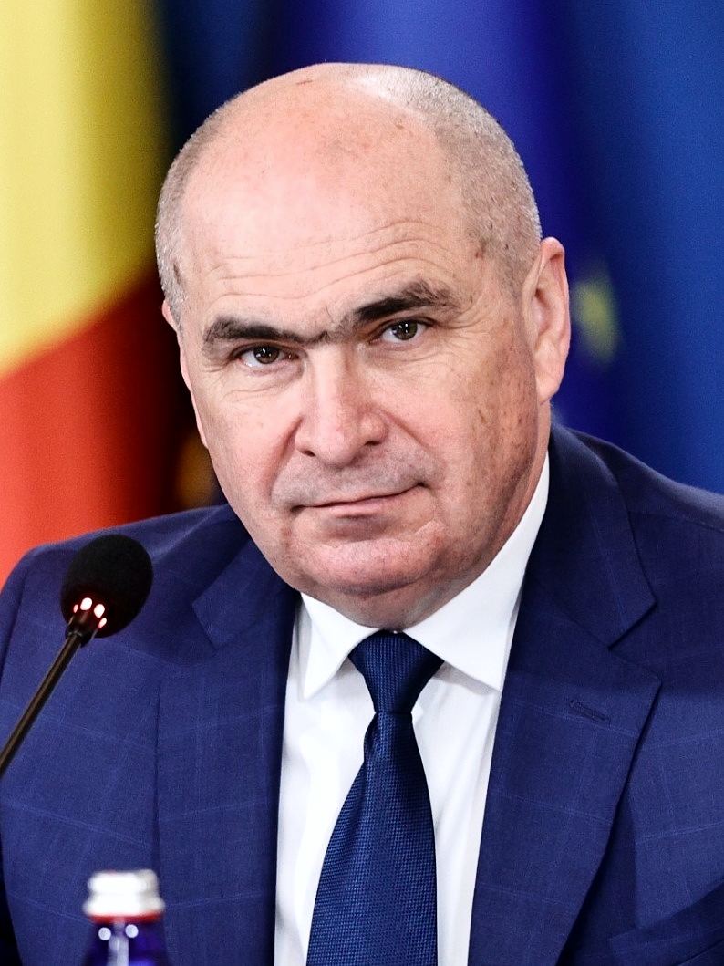 Ilie Bolojan, în costum și cravată, la o reuniune oficială, în fața drapelelor României și Uniunii Europene (fotografie de arhivă).