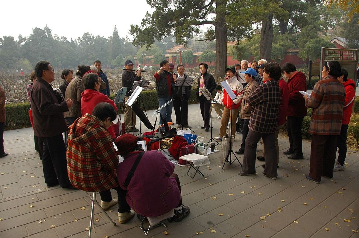 Persoane în vârstă cântând în grup, cu partituri, într-un parc din Beijing, lângă o clădire tradițională.