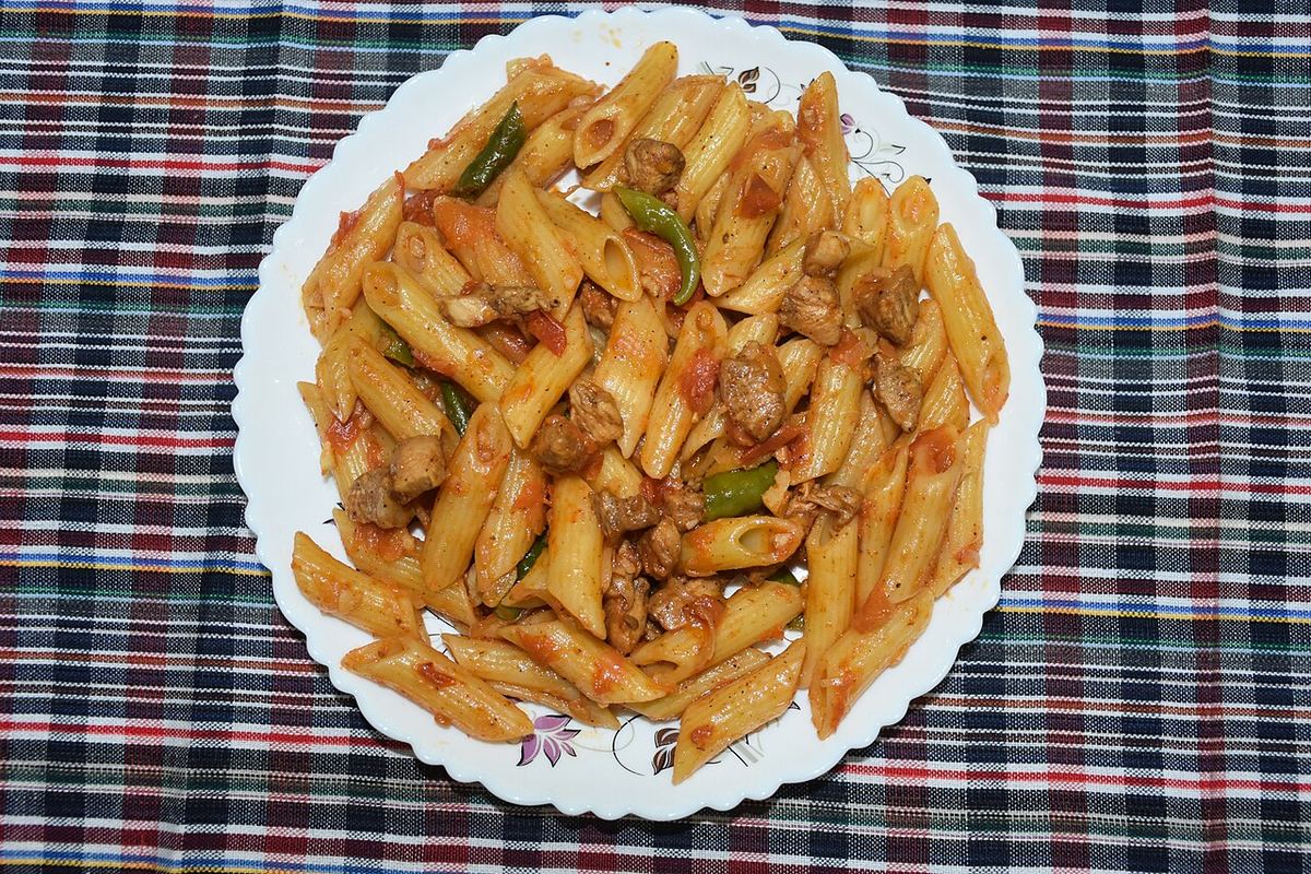 Farfale sau penne cu sos de roșii, legume verzi și bucăți de carne, servite pe o farfurie albă.