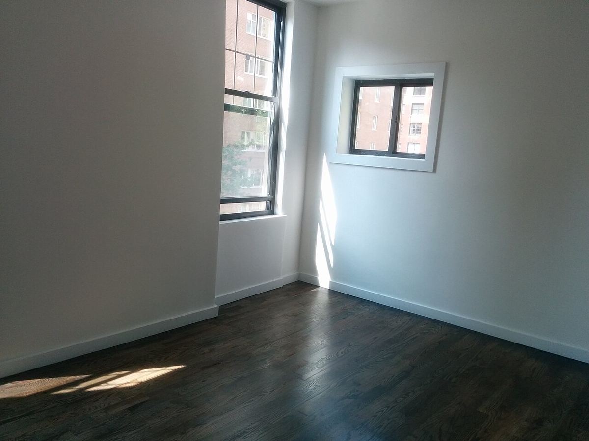 Cameră goală de apartament cu podea din lemn închis la culoare, pereți albi și două ferestre de colț care lasă lumina soarelui să intre.