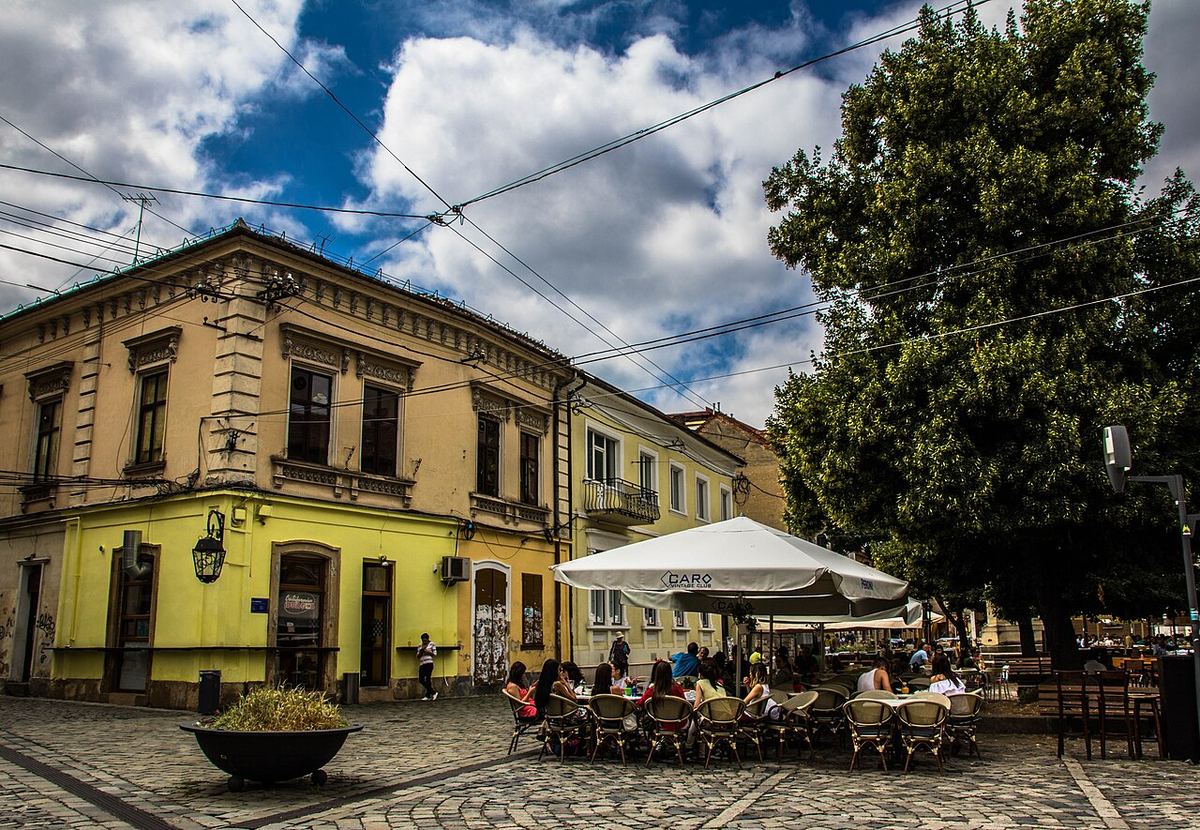 Terasă cu umbrele albe și clădire galbenă de epocă pe Piața Muzeului din Cluj-Napoca, pe timp de zi.
