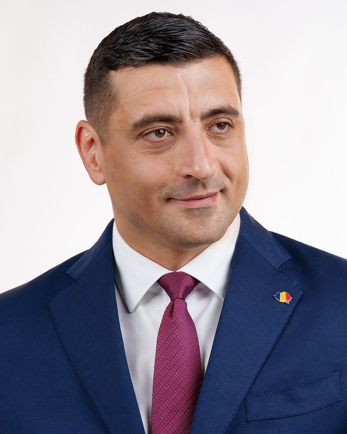 Portret în studio al lui George Simion, în costum bleumarin, cămașă albă și cravată bordo, cu insigna tricolorului pe rever.
