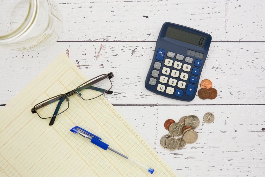 Calculator, ochelari, bloc de notițe, pix și monede pe o masă albă, ilustrație pentru gestionarea bugetului personal.