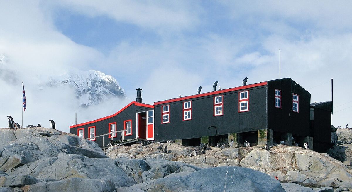 Clădirea din lemn cu acoperiș roșu de la fosta bază britanică Port Lockroy, pe o coastă stâncoasă din Antarctica, cu munți acoperiți de zăpadă.