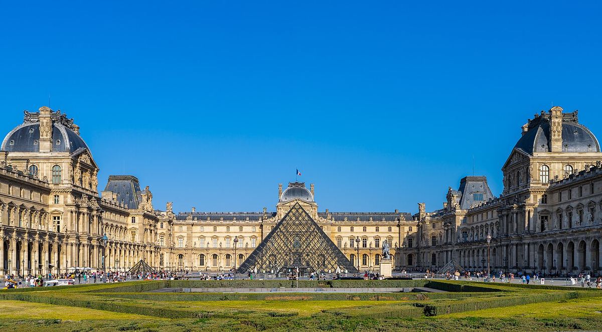 Muzeul Luvru din Paris, cu piramida de sticlă în curte, fotografiat pe timp însorit, cu vizitatori în fața clădirii.