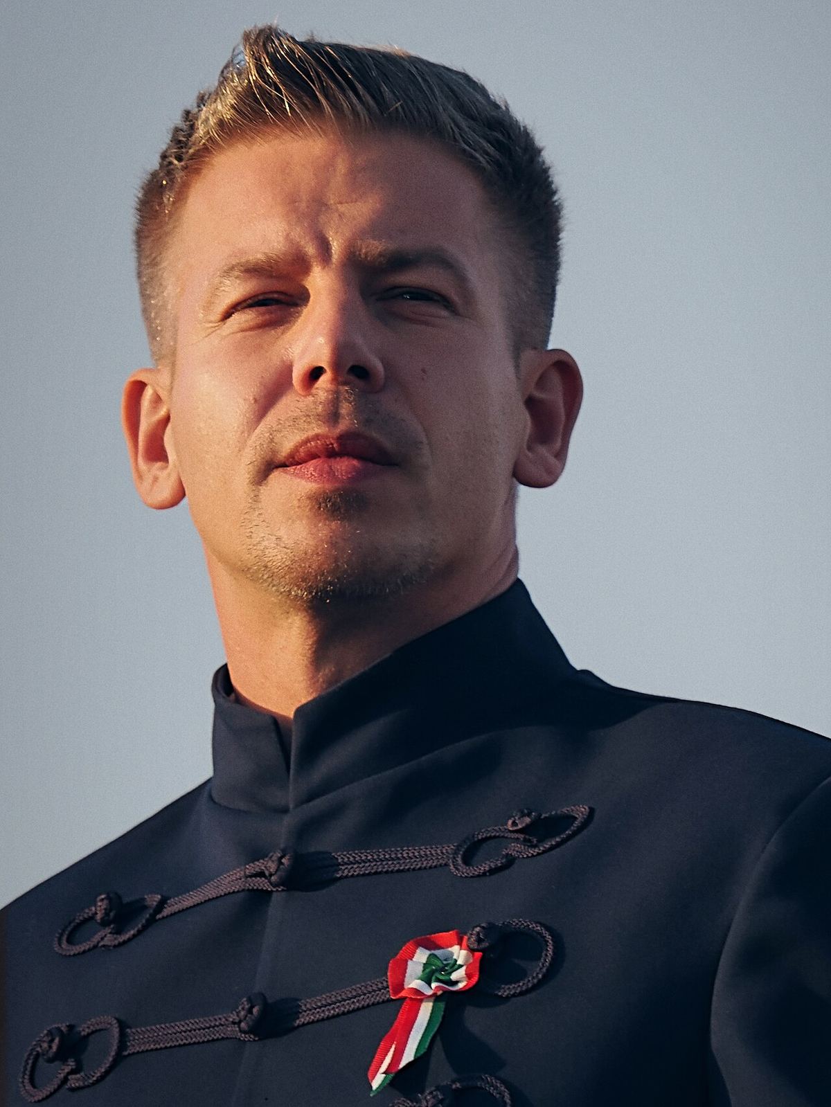 Portret al premierului Péter Magyar, într-un sacou închis la culoare, cu o cocardă tricoloră maghiară la piept, pe fundal deschis.