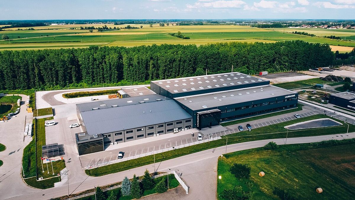 Clădire industrială modernă cu acoperiș metalic, parcare și câmpuri agricole în jur, fotografiată din aer.