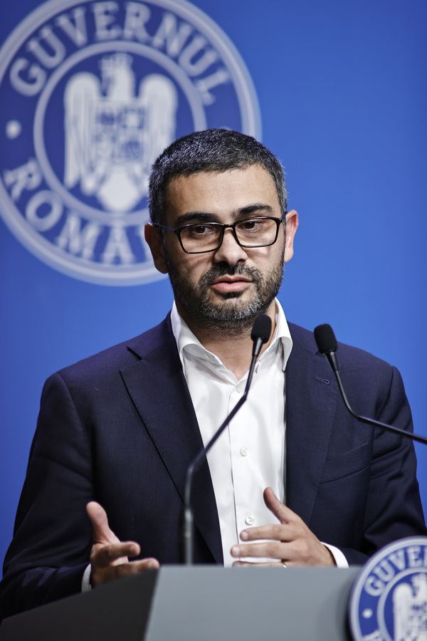 Petre Florin Manole vorbind la un pupitru de conferință de presă, în fața stemei Guvernului României, pe fundal albastru.