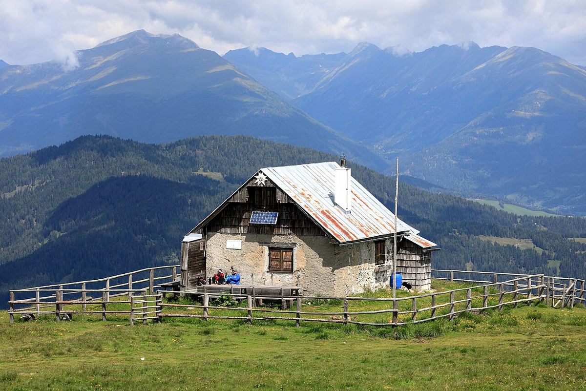Cabană alpină de munte cu acoperiș înclinat, situată într-o zonă montană cu păduri și pajiști verzi.
