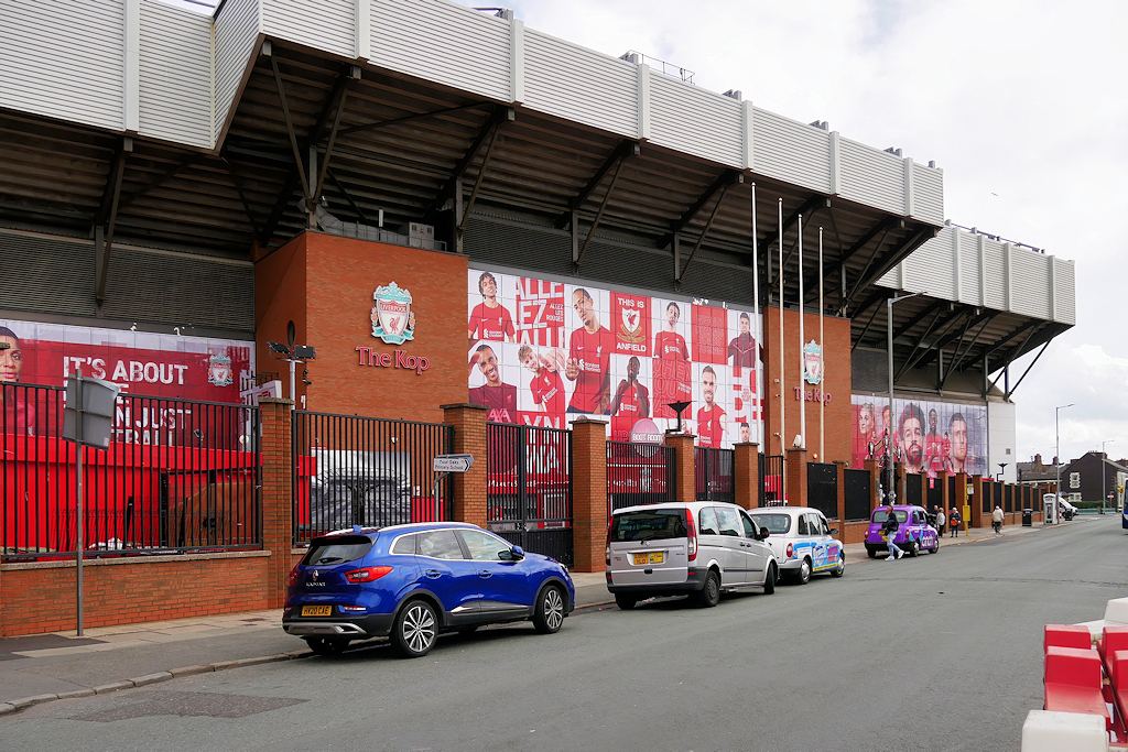 Exteriorul stadionului Anfield, zona Kop, cu banner-e ale jucătorilor Liverpool FC și mașini parcate pe stradă.
