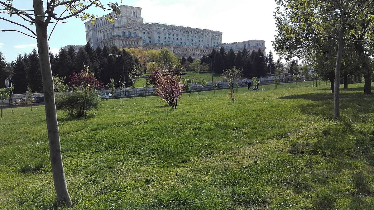 Clădire monumentală văzută din spatele unui parc cu iarbă verde și copaci, pe o zi senină, ilustrativ.