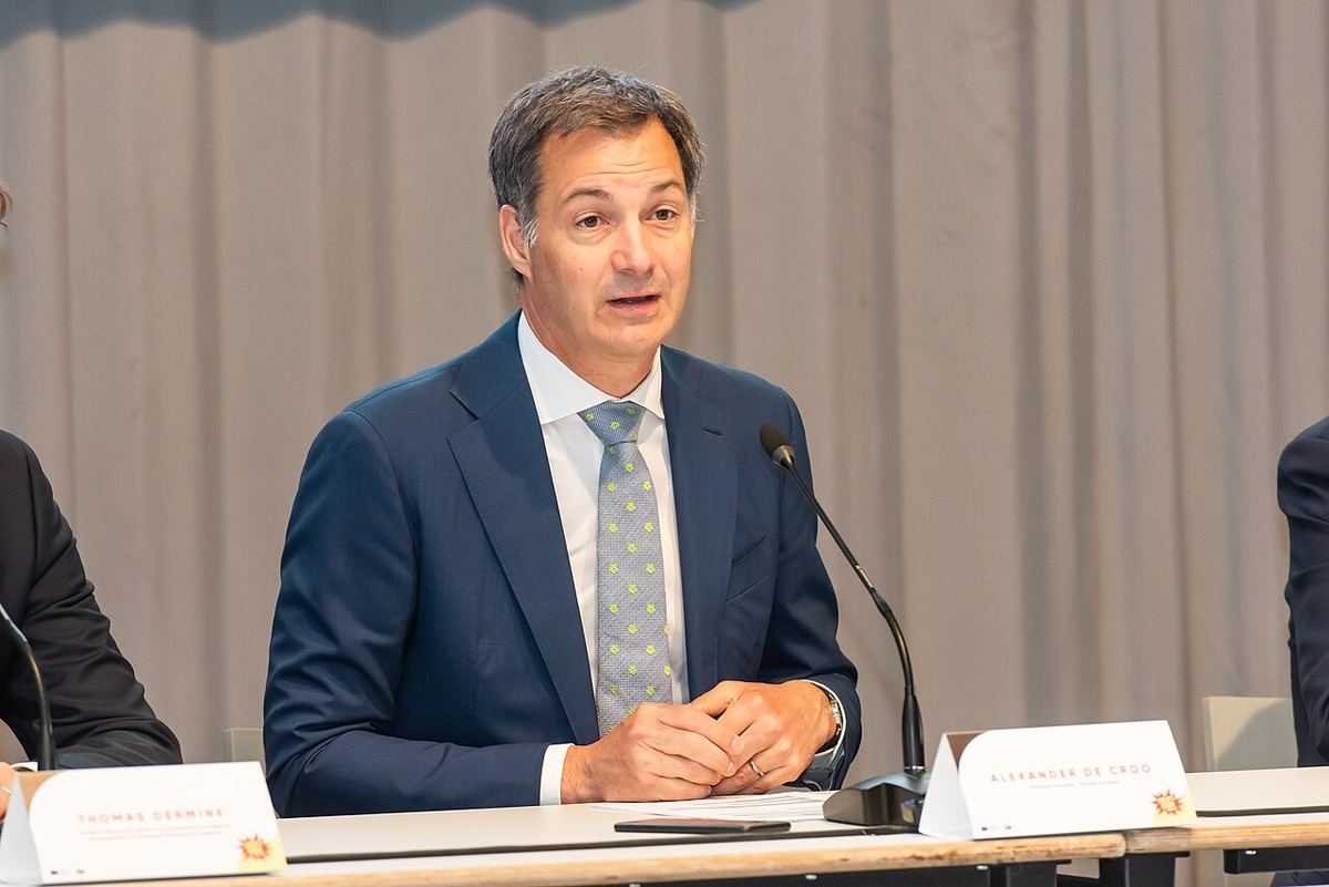 Alexander De Croo vorbind la un eveniment, așezat la o masă cu plăcuța cu numele său, în fața unei draperii gri (fotografie de arhivă).