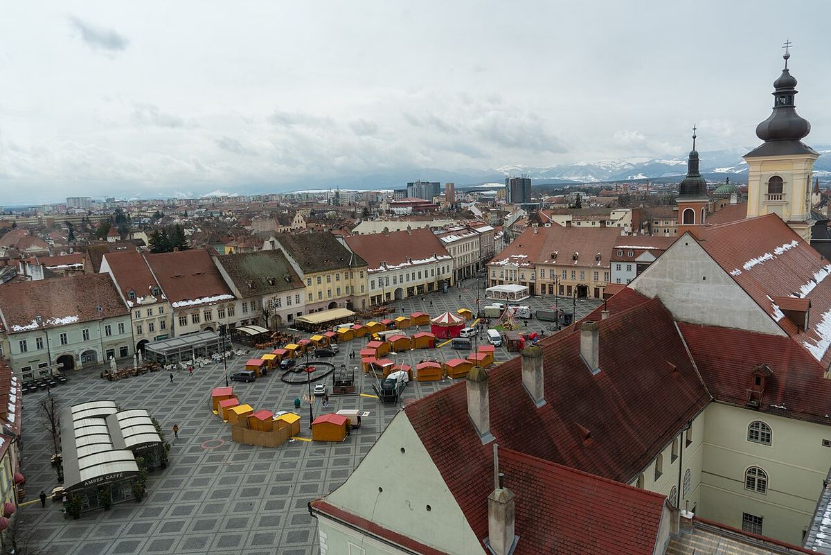 Vedere panoramică asupra Pieței Mari din Sibiu, plină de tarabe, cu turnul Bisericii Sfintei Treimi și Carpații acoperiți de zăpadă la orizont.