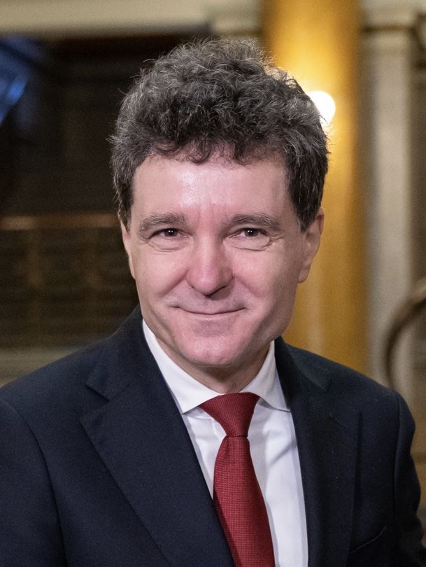 Portret al președintelui Nicușor Dan, în costum bleumarin și cămașă albă, cu cravată roșie, zâmbind.