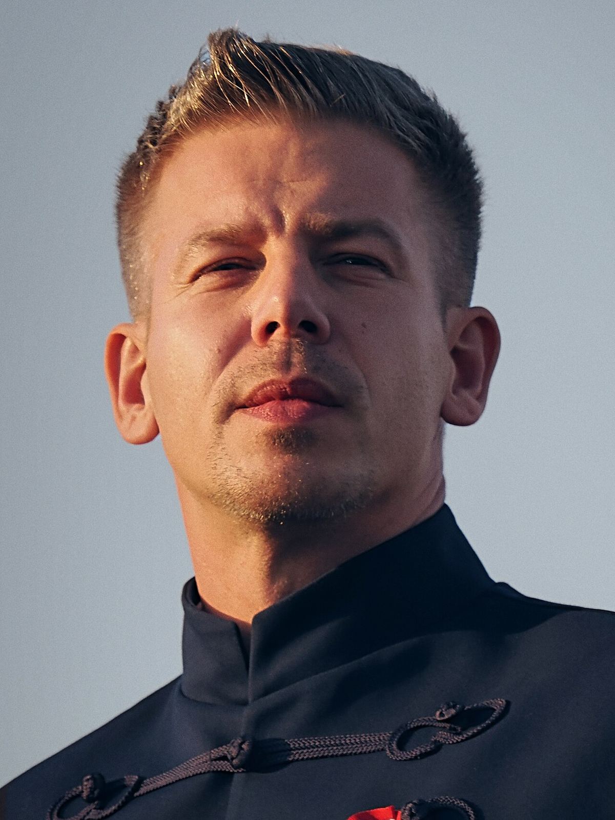 Portret al lui Péter Magyar, cu păr scurt și jachetă neagră cu broderie, fotografiat în aer liber pe fundal deschis.