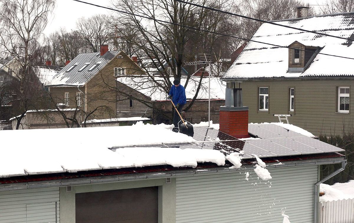 Un bărbat în geacă albastră curăță zăpada de pe panourile fotovoltaice de pe un acoperiș, în Tartu, Estonia.