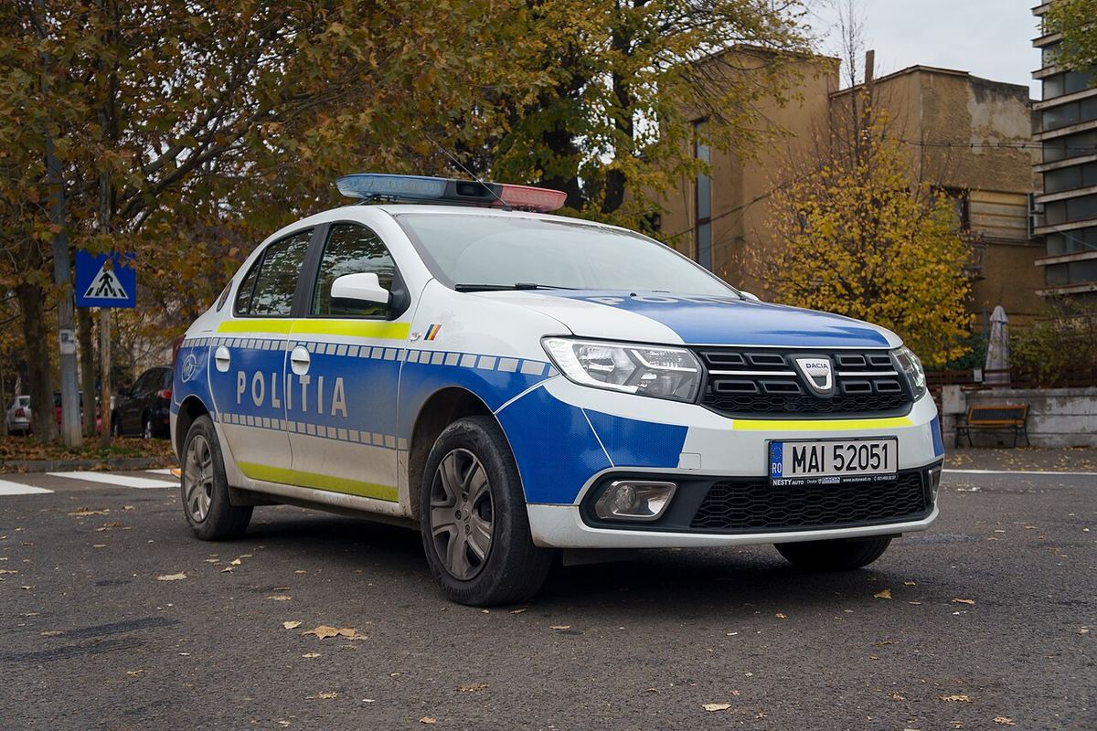 Autoturism Dacia Logan al Poliției Române, cu livrea albastră și galbenă, parcat pe o stradă cu frunze uscate.