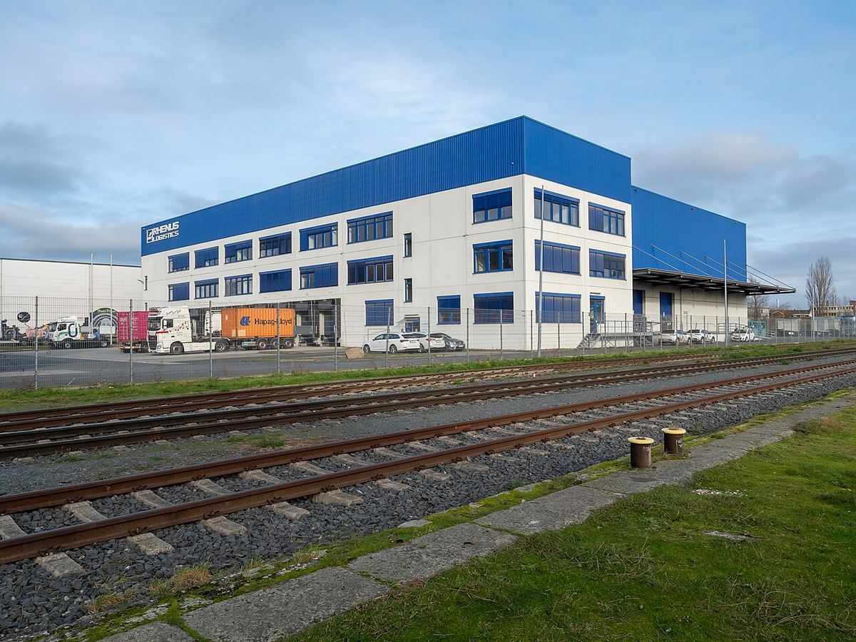 Depozit logistic cu fațadă albă și albastră, camioane în curte și linii de cale ferată în prim-plan (Frankfurt, Germania).
