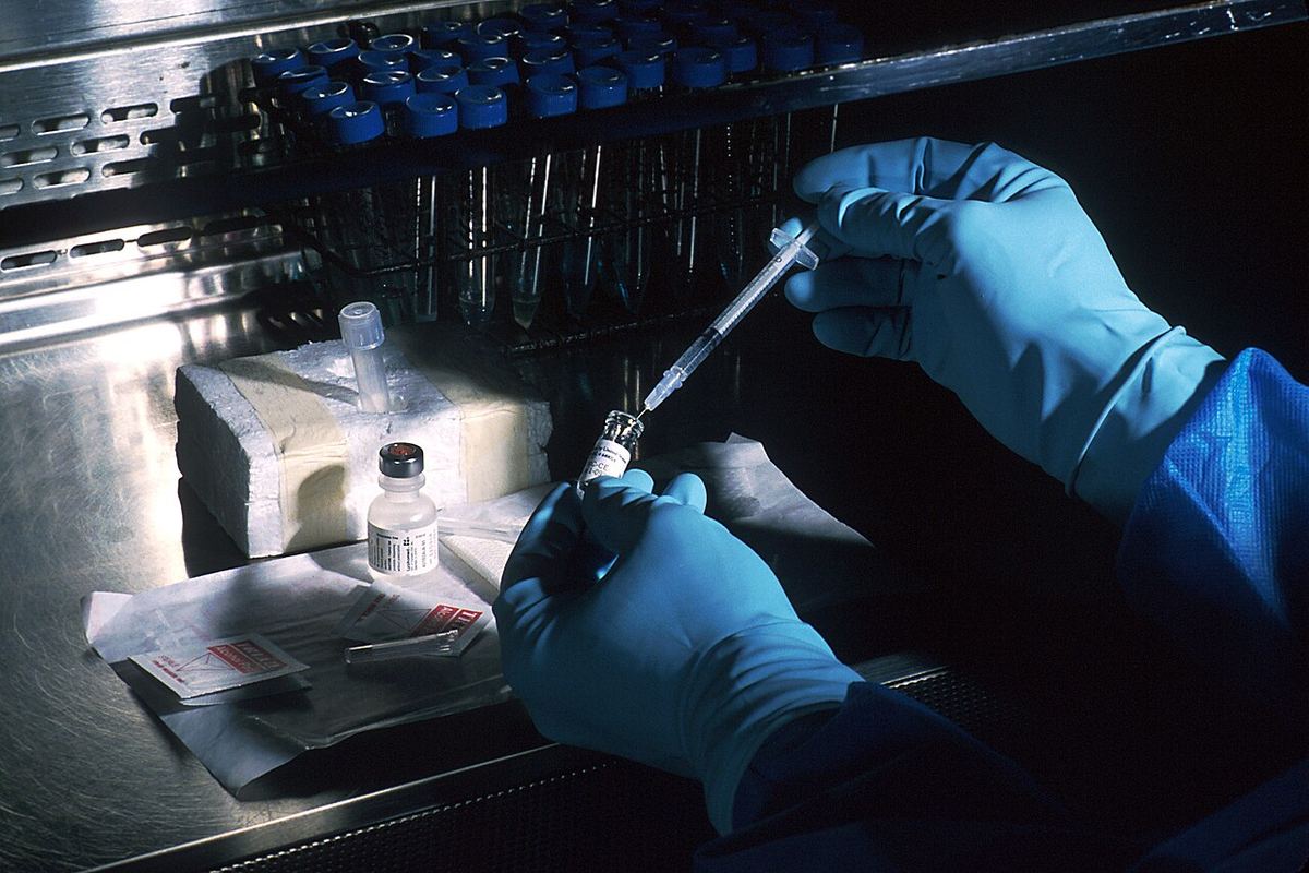 Mâini cu mănuși albastre pregătind o seringă și un flacon cu vaccin într-un laborator medical. Imagine ilustrativă.