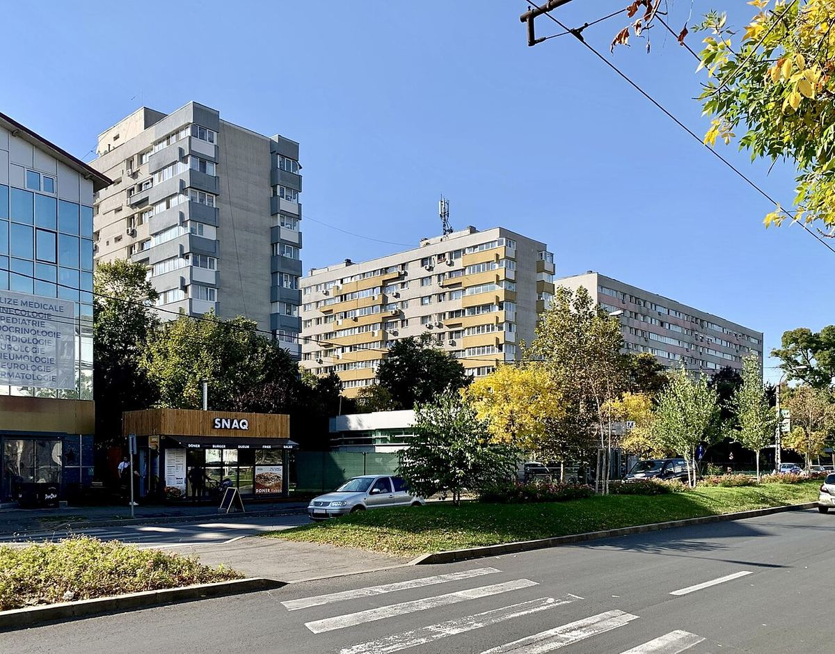 Blocuri de locuințe din anii 1960 pe un bulevard din București, cu o mașină albă parcată și copaci în prim-plan.