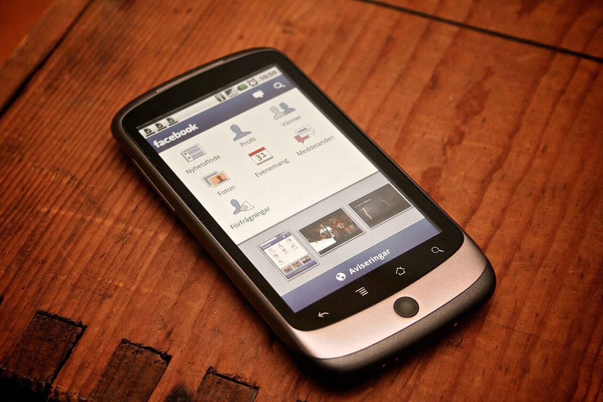 Telefon mobil cu aplicația Facebook pe ecran, așezat pe o suprafață de lemn.
