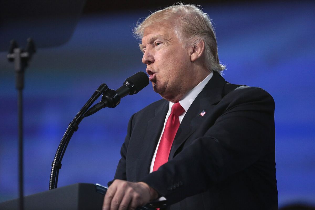 Donald Trump vorbind la microfon, la un pupitru, în costum închis și cravată roșie.