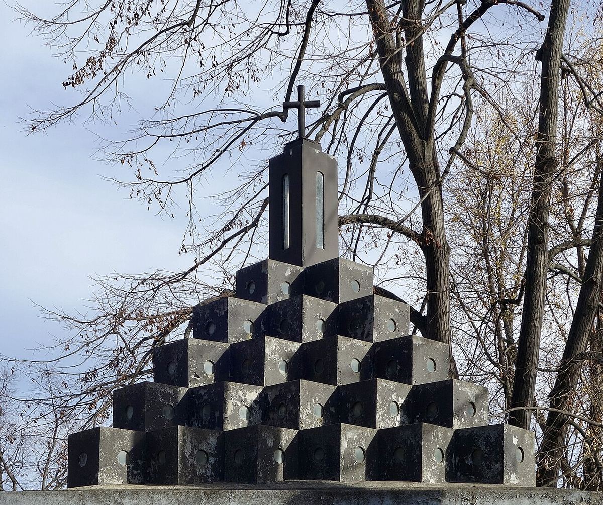 Mormânt cu monument funerar din piatră într-un cimitir, cu cruce și inscripții, sub lumina zilei.