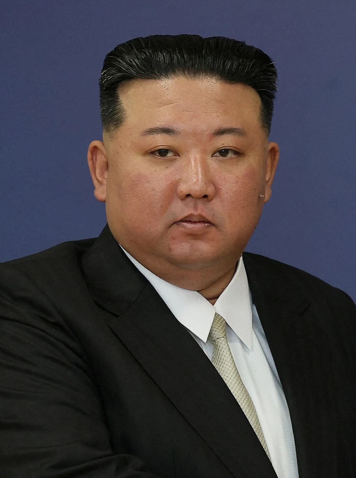 Fotografie de arhivă cu Kim Jong-un, în costum închis, cămașă albă și cravată, pe fundal albastru.
