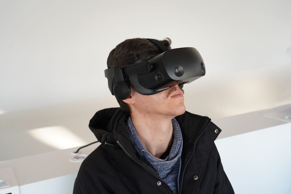 Persoană cu cască de realitate virtuală neagră, privind ușor în sus, îmbrăcată în jachetă închisă și pulover albastru, pe fundal alb.
