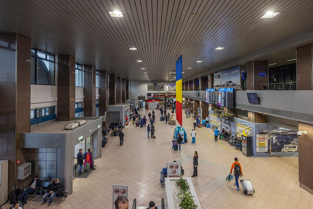 Interiorul terminalului Aeroportului Henri Coandă din București, cu pardoseli, plafon din lamele și iluminat încastrat, și pasageri în hol.