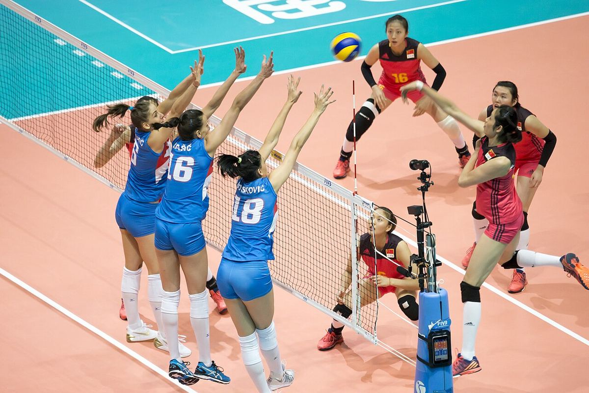 Trei jucătoare de volei fac un bloc triplu la fileu, într-un meci internațional, ilustrativ pentru jocul de volei feminin.