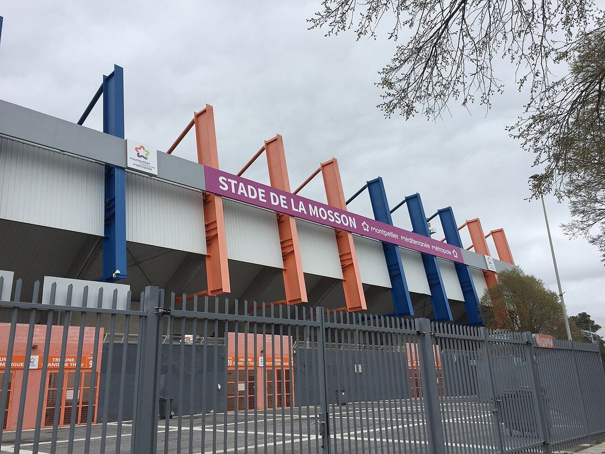 Exteriorul Stadionului Stade de la Mosson din Montpellier, cu tribune, stâlpi colorați și gard metalic, pe un cer înnorat.