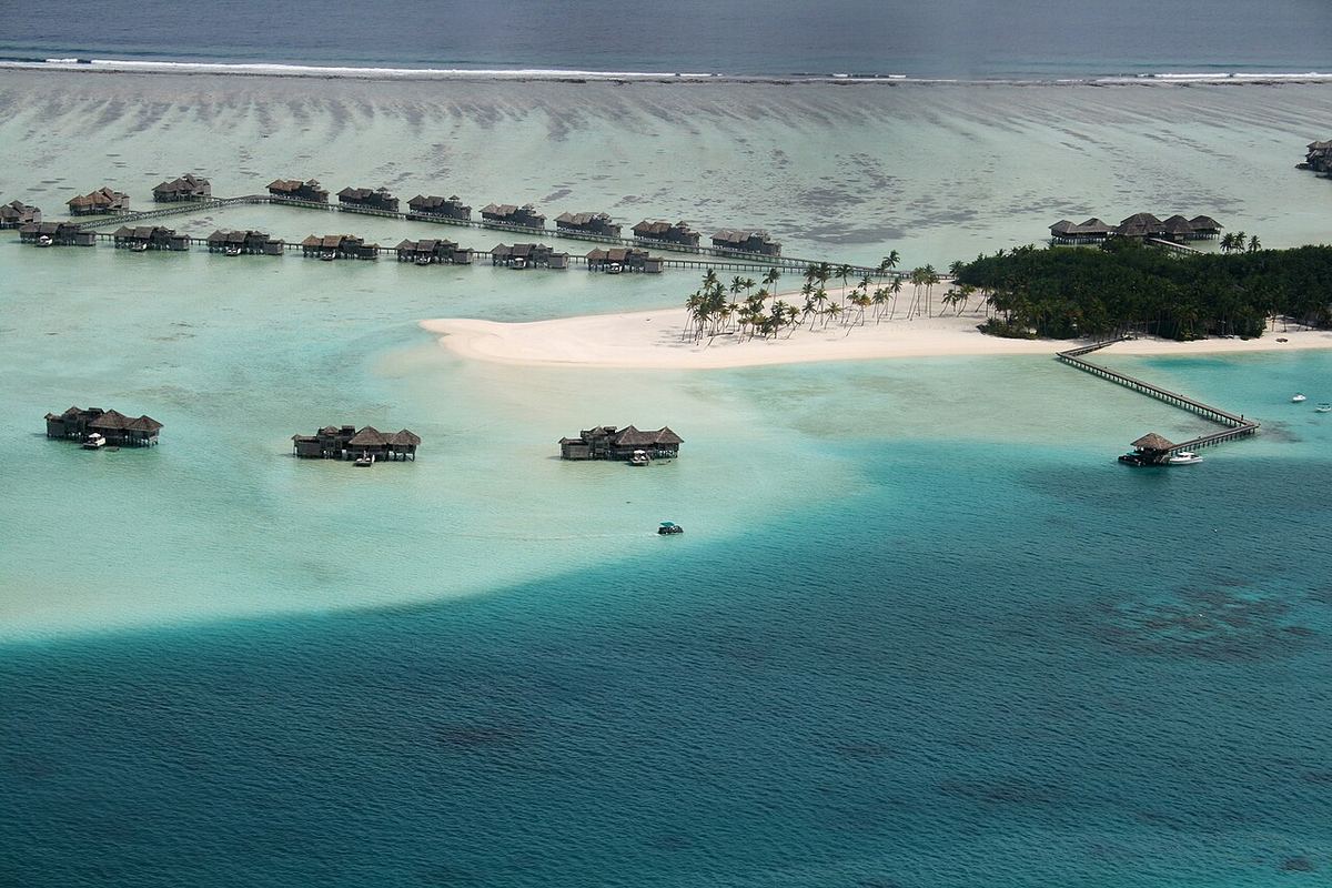 Vedere aeriană a unei insule joase din Maldive, cu plaje de nisip, vile pe apă și apă turcoaz în jurul recifului de corali.
