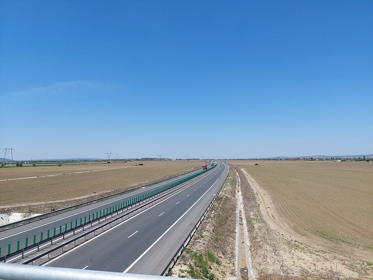 Autostradă modernă în România, cu două benzi pe sens și parapet verde, traversând câmpuri agricole sub cerul senin.