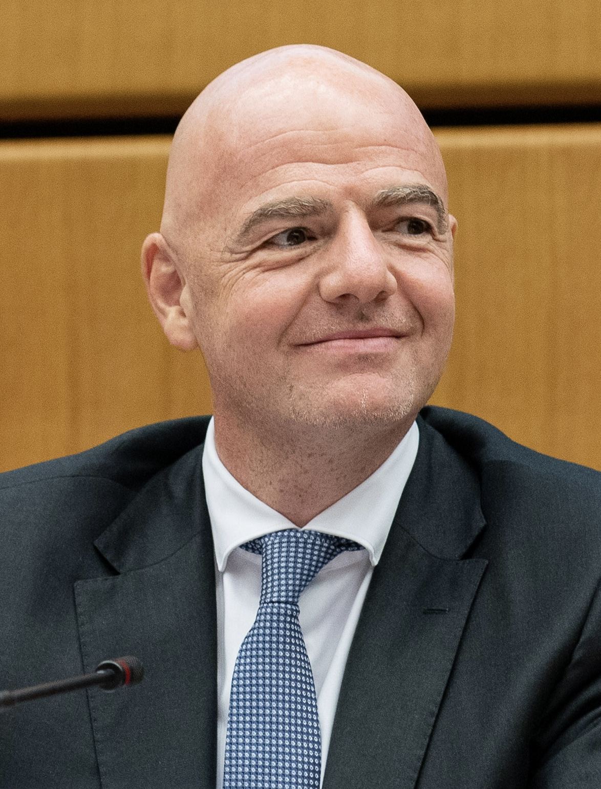 Gianni Infantino, în costum închis, cămașă albă și cravată albastră, la o masă cu microfon. Fotografie de arhivă.