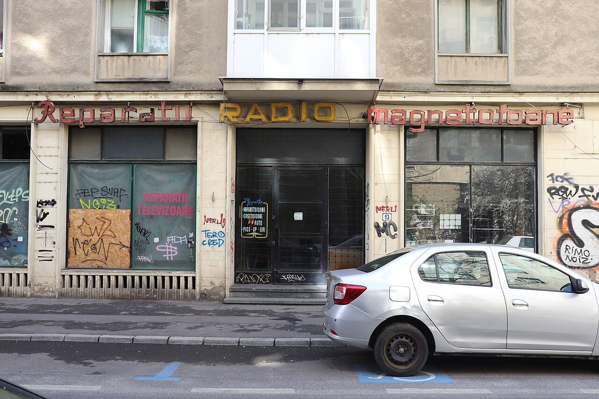 Fațada unei clădiri din București cu vitrine închise, afișe și graffiti, pe o stradă cu mașini parcate.