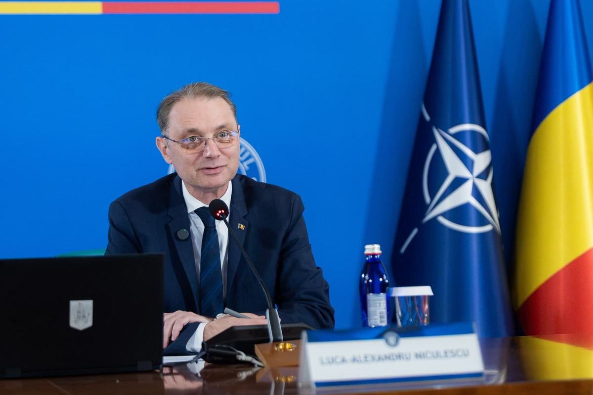 Premierul Luca Niculescu la masa ședinței, cu steagurile României, UE și NATO în spate