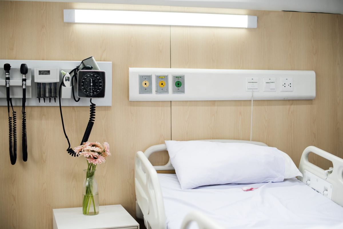 Pat de spital făcut și nefolosit, lângă un vas cu flori, cu aparate medicale pe perete, într-o salon de spital.
