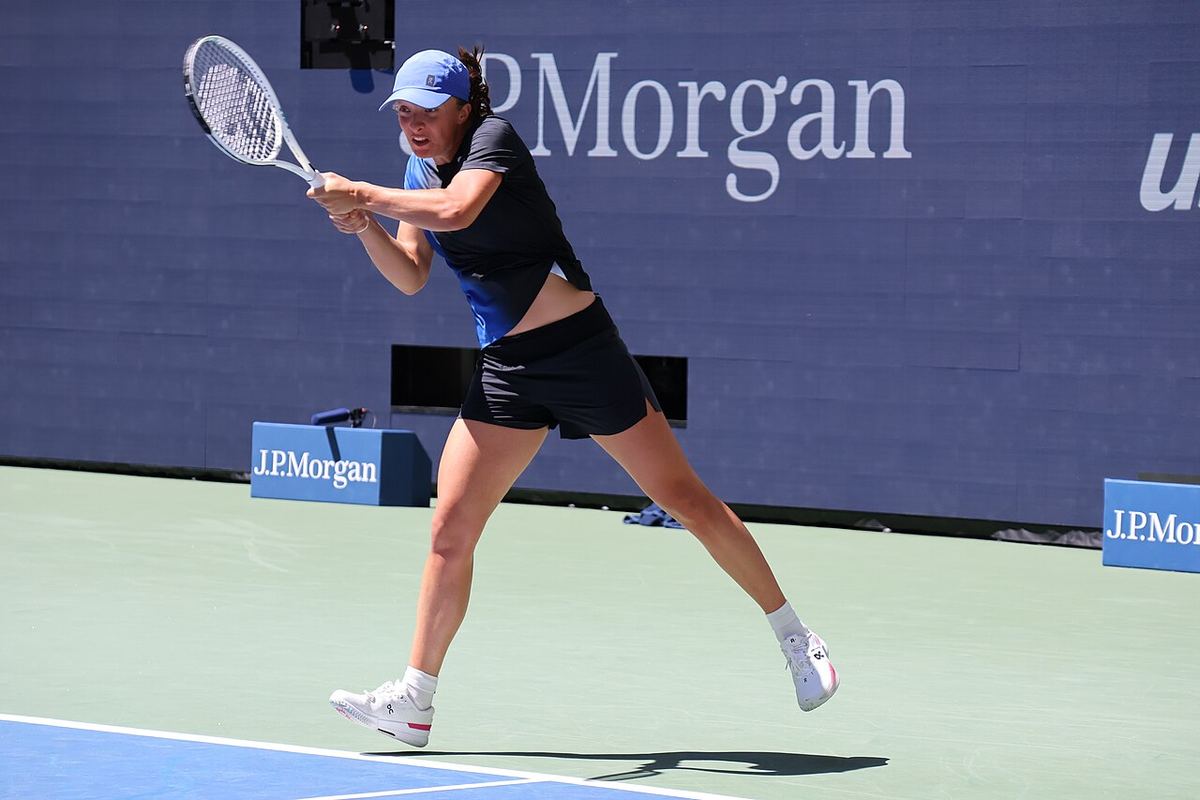 Iga Świątek, cu șapcă albastră, lovind cu revers pe un teren de tenis dur, în fața unui panou J.P. Morgan.