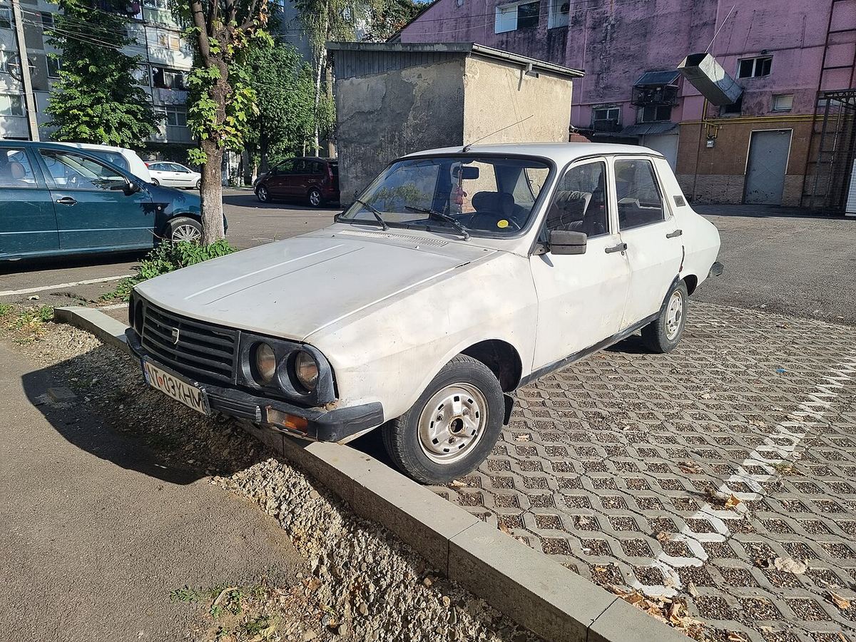 Dacia 1310 albă, parcată pe un trotuar dintr-un oraș din România, cu caroseria boxy caracteristică modelului.