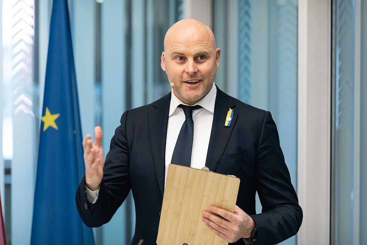 Bărbat chel, în costum bleumarin și cămașă albă, gesticulând și ținând o planșetă de lemn; în fundal, steagul UE.