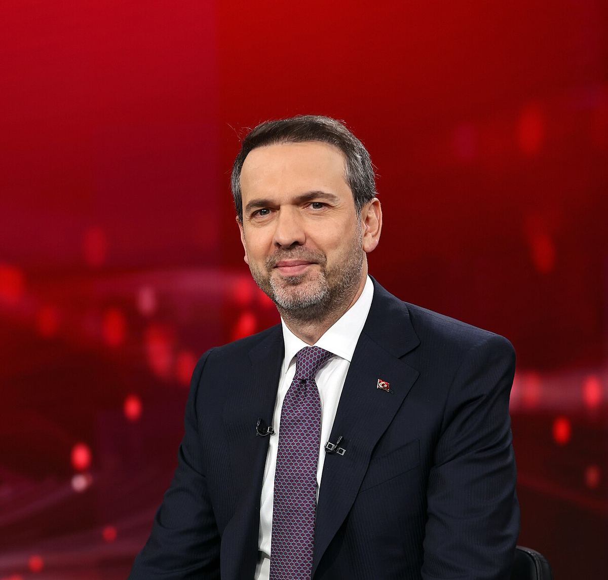 Alparslan Bayraktar, ministrul turc al Energiei, în costum bleumarin și cravată mov, pe fundal roșu, în studio.